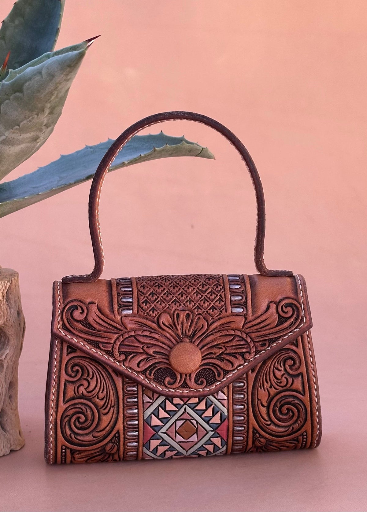 Handcrafted leather micro handbag for cards and cash with intricate tooled patterns next to an agave on a pink background
