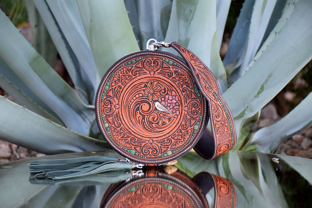 A round tooled leather crossbody bag with intricate brown and turquoise floral engravings and a small bird motif, resting on a reflective surface with agave leaves in the background.