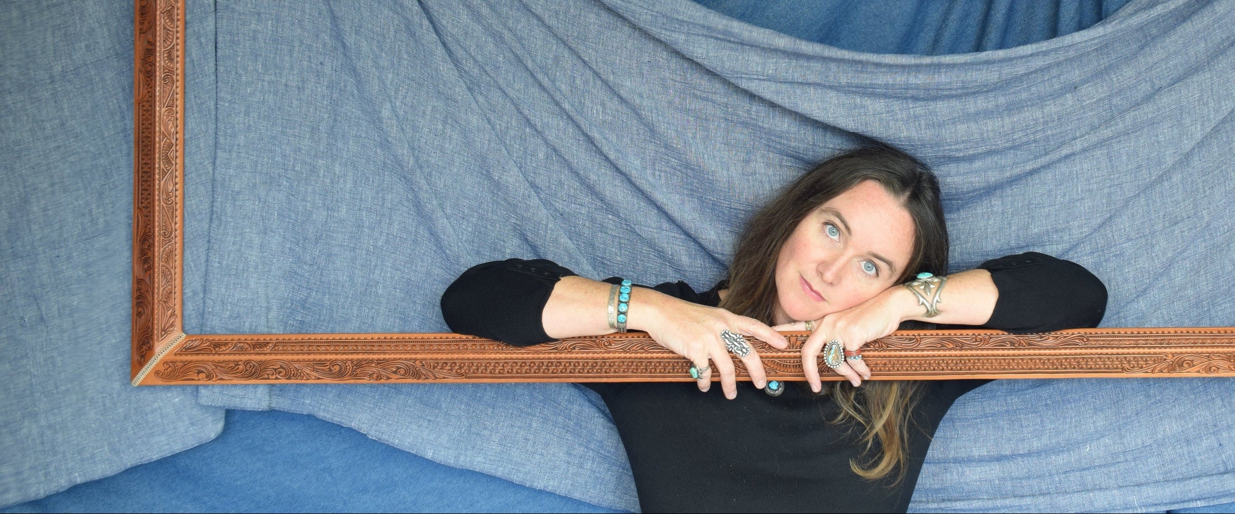 A woman, the artist, with long dark hair peeks through a large rectangular leather picture frame she custom made hanging in front of draped blue fabric. She is wearing a black long-sleeve top and has bracelets on her wrists.