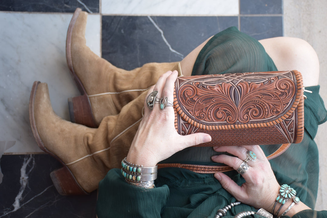 A close-up lifestyle shot of a person holding a brown tooled leather clutch bag, wearing a dark green dress, tan suede cowboy boots, and silver jewelry with turquoise accents, seated on a black and white checkered floor.