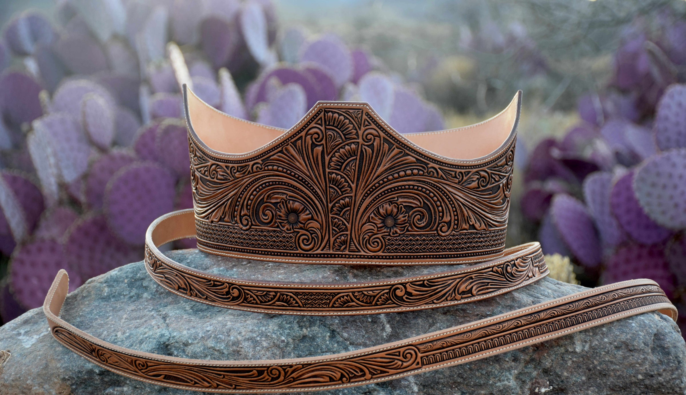 A tooled leather corset belt with matching narrow fashion belt, displayed on a rock with a blurred background of purple cacti. The belts features intricate floral and scroll engravings in dark brown on a natural veg-tan leather base.