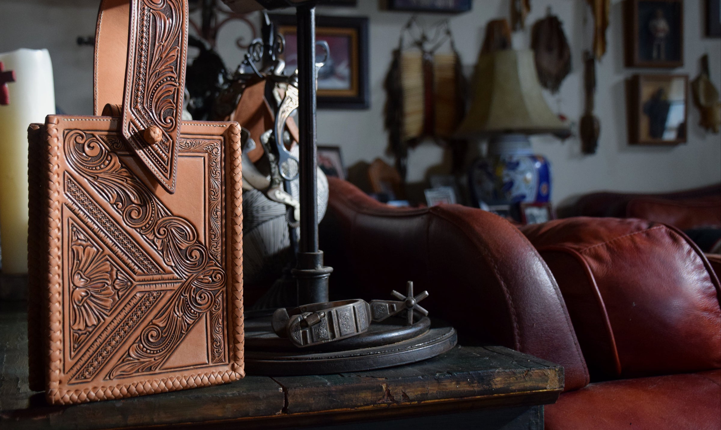 A brown leather handbag with intricate hand-tooled western-style floral and geometric patterns, standing on a rustic wooden table in a dimly lit, cozy living room next to handmade spurs and bits