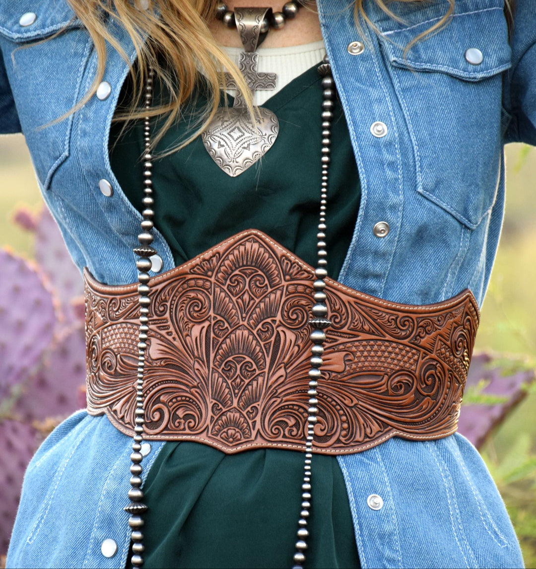 A close-up of a woman wearing a wide, heavily tooled brown leather western waist belt over a dark green top and blue denim jacket, with a blurred outdoor background featuring cacti.
