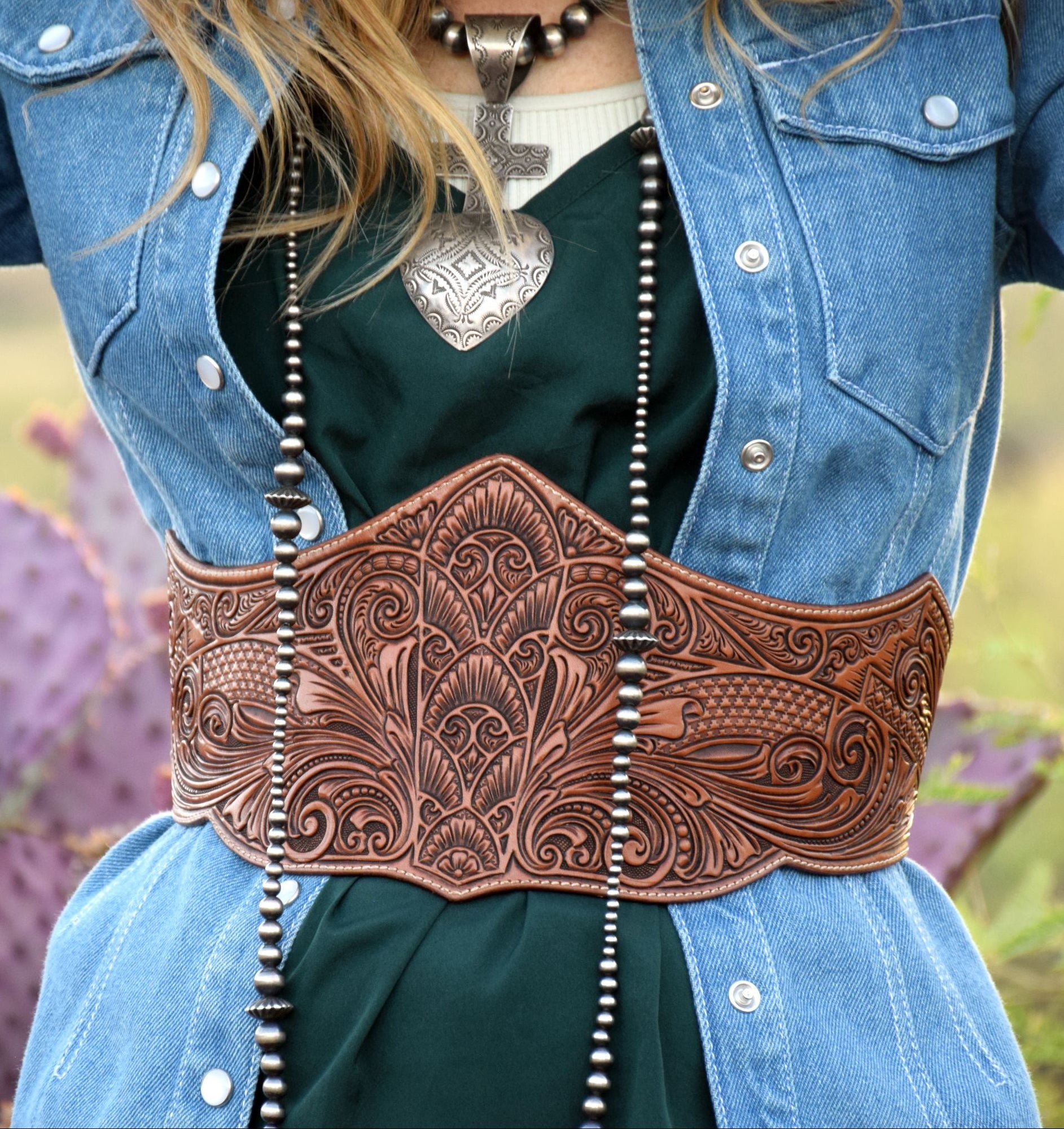 A close-up of a woman wearing a wide, heavily tooled brown leather western waist belt over a dark green top and blue denim jacket, with a blurred outdoor background featuring cacti.