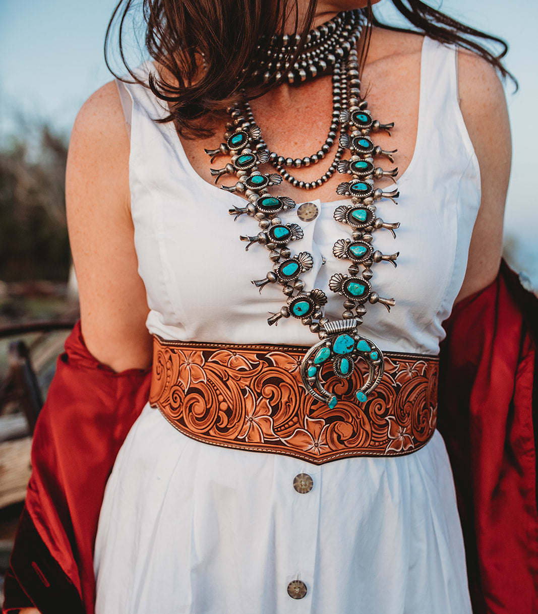 Closeup of woman in white dress wearing squash blossom and silver necklaces and a tooled leather waist cincher belt.  The belt is carved with intricate scrollwork and flower motifs.