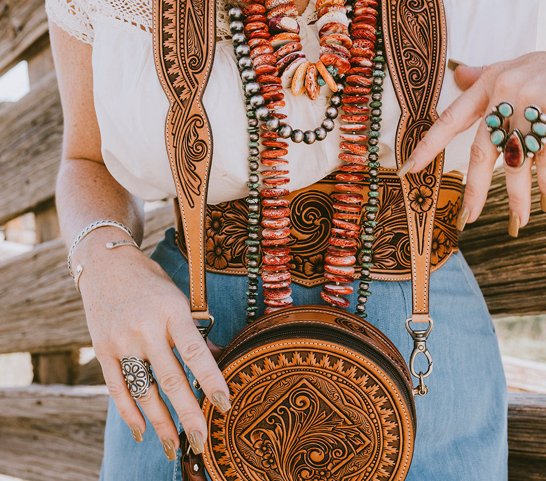 A close-up lifestyle shot of a woman wearing a white lace top and blue denim skirt, holding a round, hand-tooled brown leather crossbody bag with a tassel. The person is also wearing multiple southwestern-style beaded necklaces and rings and a tooled leather waist belt, standing in front of a rustic wooden fence. 