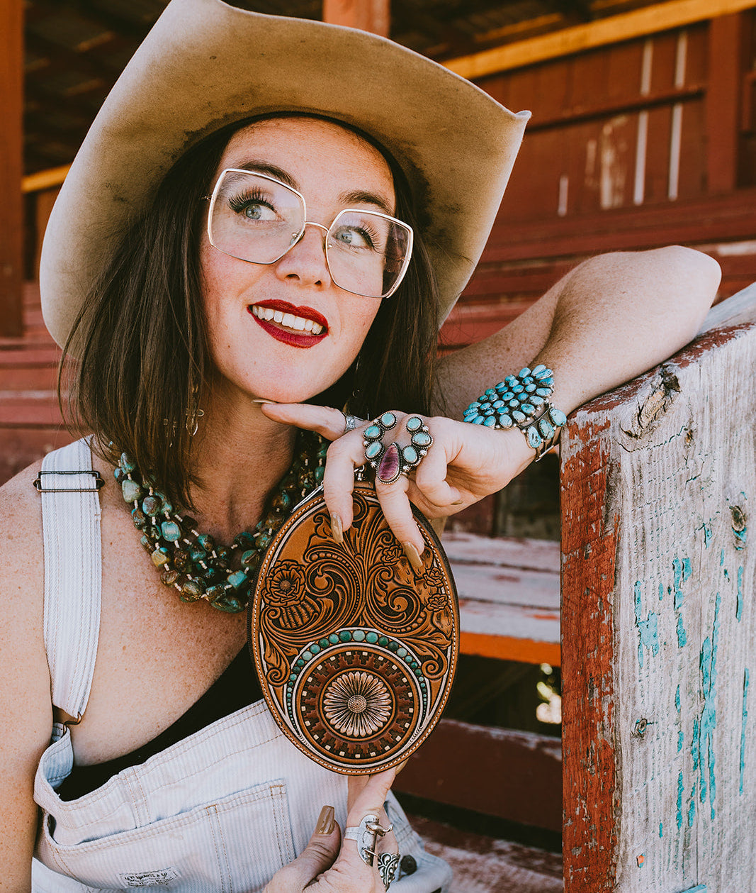 A woman in a cowboy hat and denim overalls holds a round, hand-carved leather clutch with turquoise accents, posed on rustic wooden rodeo stand.