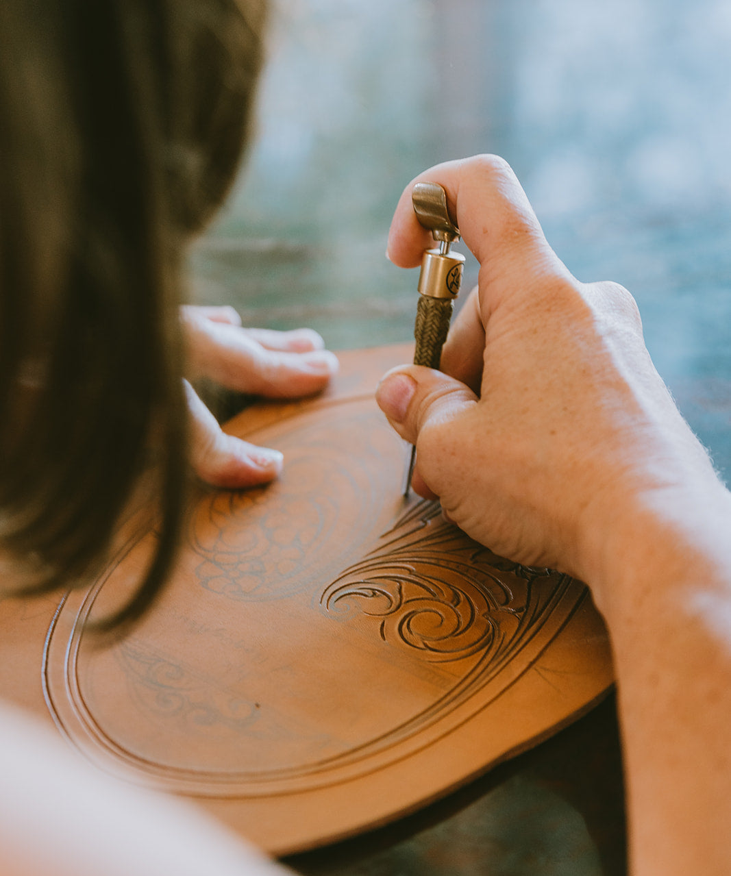 A close-up lifestyle shot of a person carving an ornate scroll pattern into a curved piece of natural leather using a swivel knife hand tool.