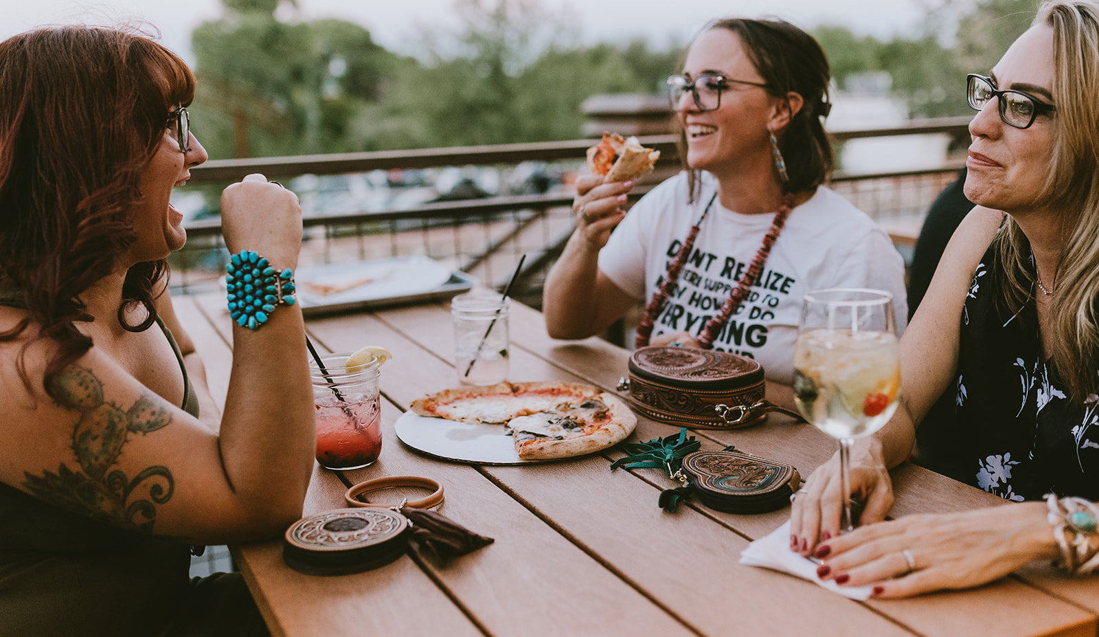 Three women laughing and eating pizza on a wooden table at an outdoor patio, with drinks and various tooled leather purses on the table.